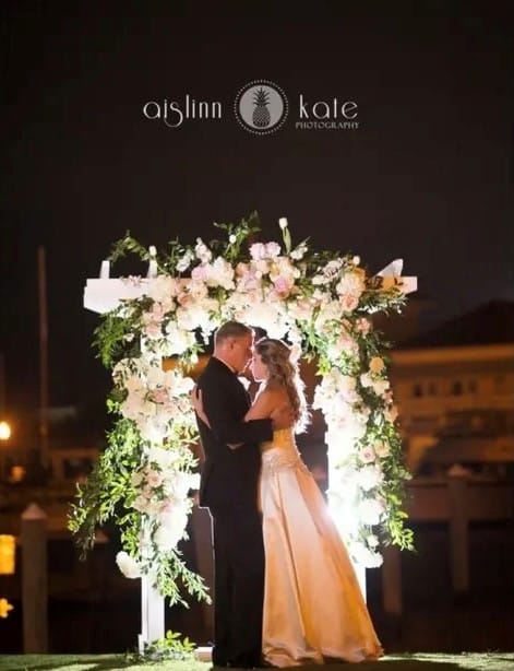 Wedding Bride and Groom at night embracing in front of their Wedding Ceremony Arbor beautifully decorated with pink and white florals with greenery at Palafox Wharf Waterfront in Pensacola, Florida, wedding venue pensacola