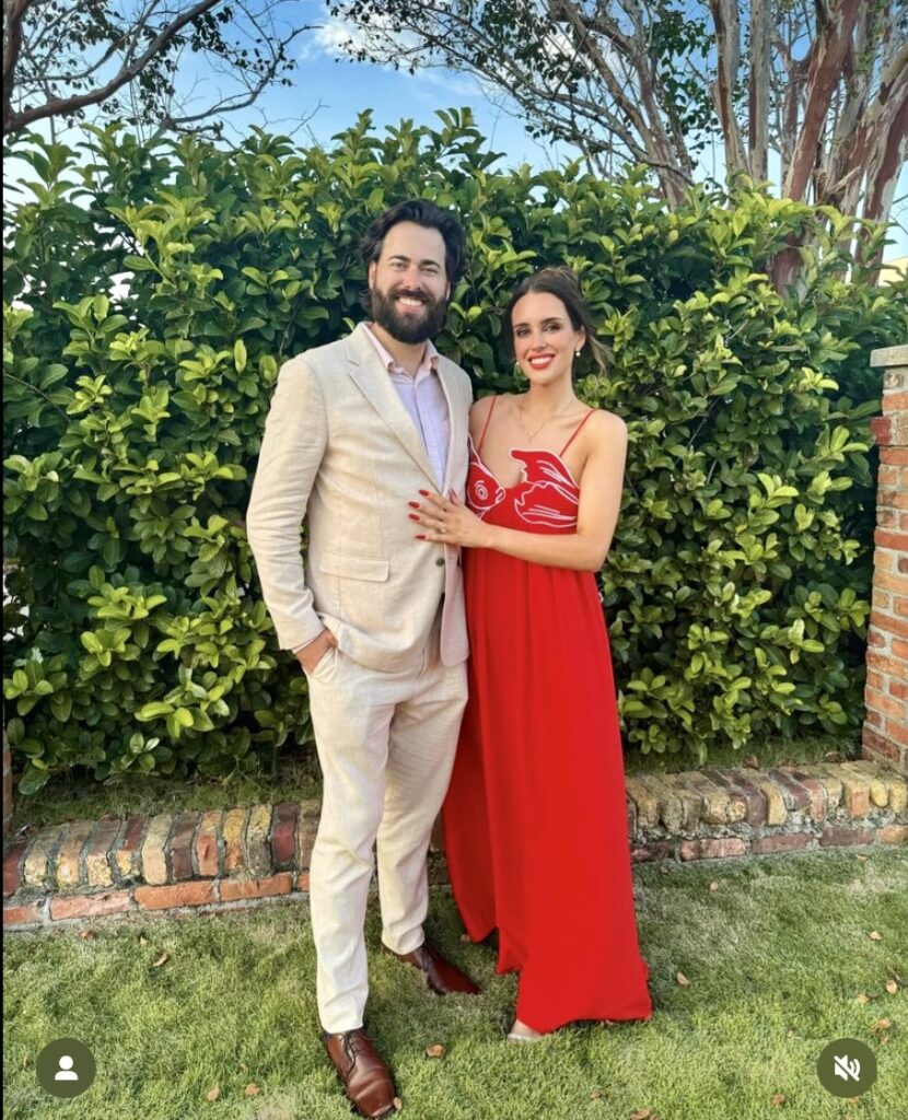 Bridesmaid and her Husband at Palafox Wharf Waterfront of a Bridesmaid in striking red gown standing in front of lush green hedge on the waterfront lawn https://617palafoxwharf.com/ https://www.thehillcrestestate.com/