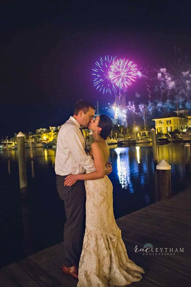 Couple shares a romantic kiss on the Palafox Wharf Waterfront dock, with fireworks illuminating the waterfront near downtown Pensacola and Destin https://www.belltowerfalls.com/ https://butlerscourtyard.com/