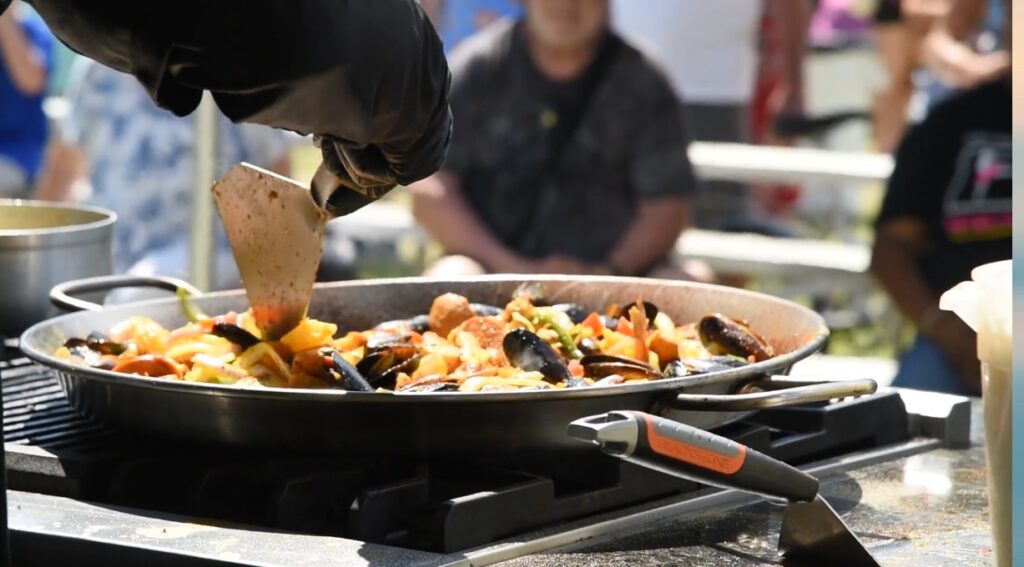 A chef serves a colorful seafood paella at an outdoor event, near wedding venues Pensacola FL, with people enjoying the vibrant atmosphere.