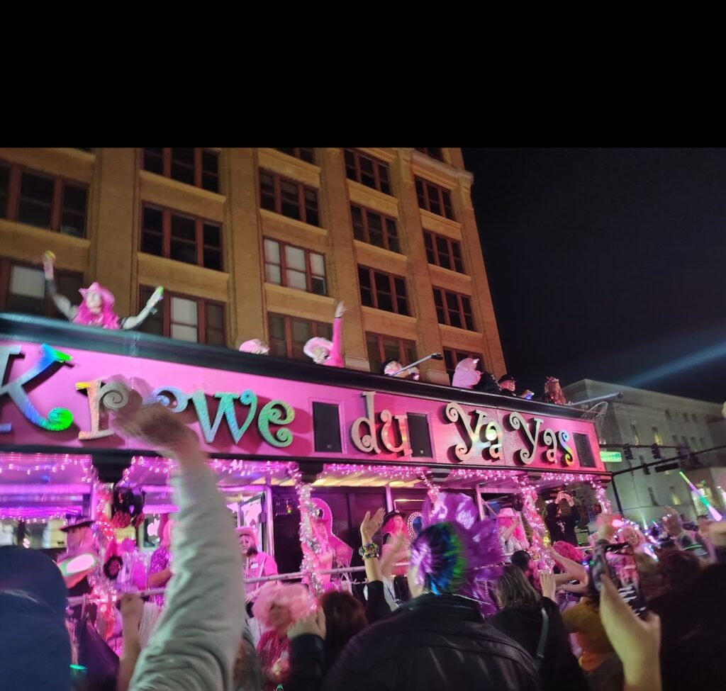 A vibrant parade float with costumed revelers, "Krewe du YaYas," passes close to a Pensacola wedding venue, with a cheering crowd under night lights.