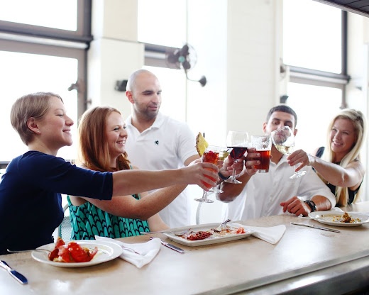 Friends toast with drinks after viewing a Pensacola wedding venue, at a cooking night surrounded by elegant decor, plates of food, and large windows offering natural light.