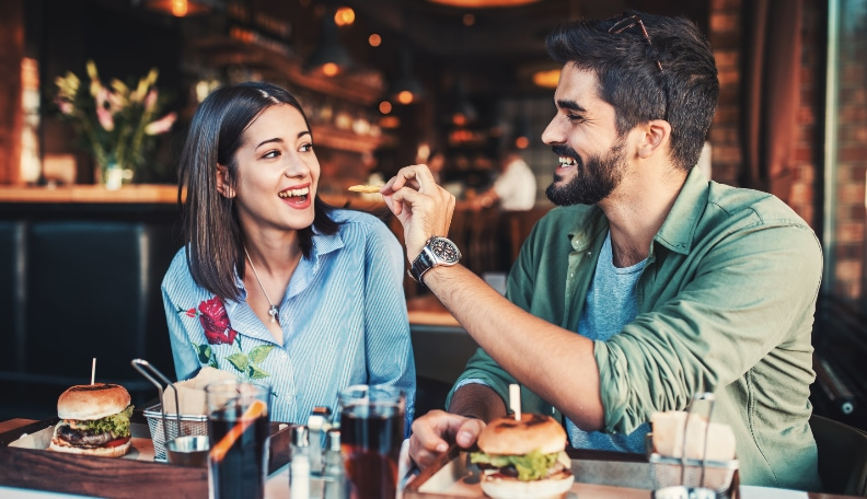 Dating couple eating hamburgers all the way and he is offering her a chip.  She is a brunette dressed in blue and he is a brunette dressed in blue and green.  They are at a restaurant.  Great First-Date Conversation Starters.
https://www.hightowerfalls.com/ 
https://617palafoxwharf.com/