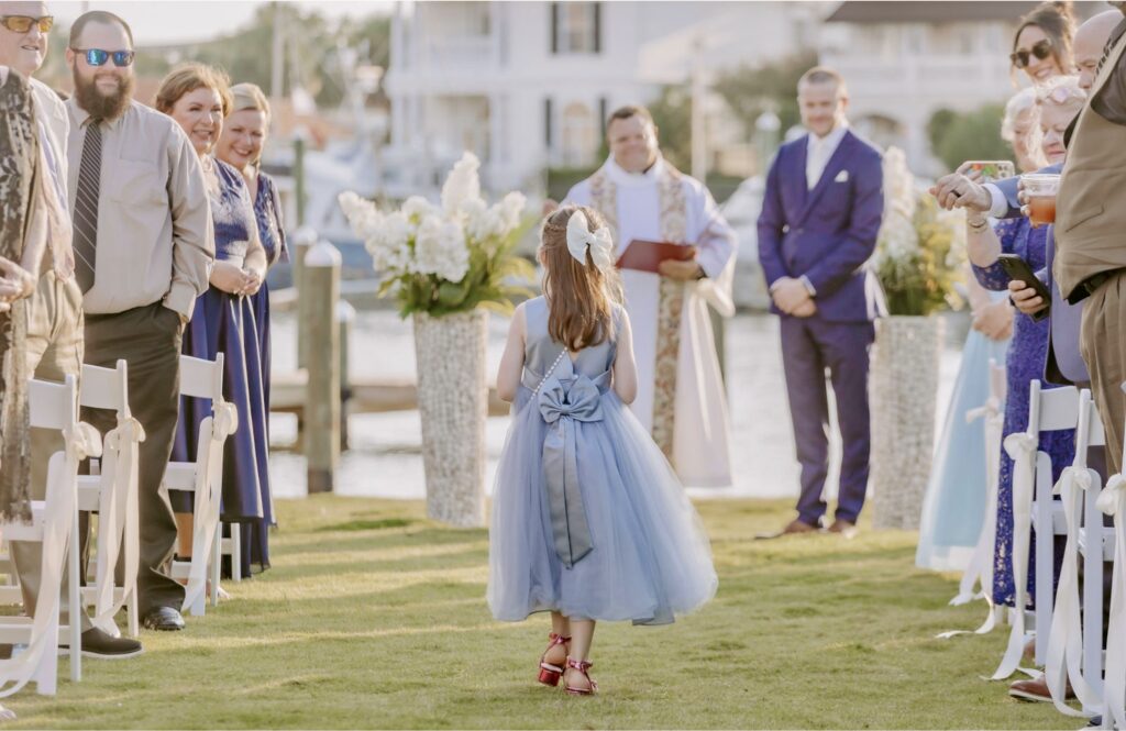 Flower girl in blue dress walking up the aisle with groom waiting with waterfront in the background at Palafox Wharf Waterfront near Pensacola Florida Events