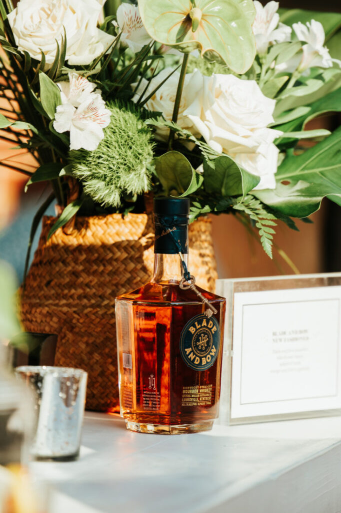 Garden + Gun’s Blade and Bow Whiskey displayed on the deck cocktail bar with floral arrangement in white and green in the back ground with a small votive in the foreground. Garden & Gun’s Scenic Downtown Cocktail Party at Palafox Wharf Waterfront