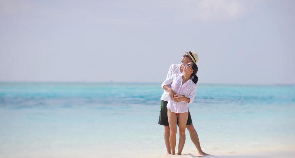 Luxury honeymoon on clear water beach.  Couple in bathing suits with male wearing a hat.