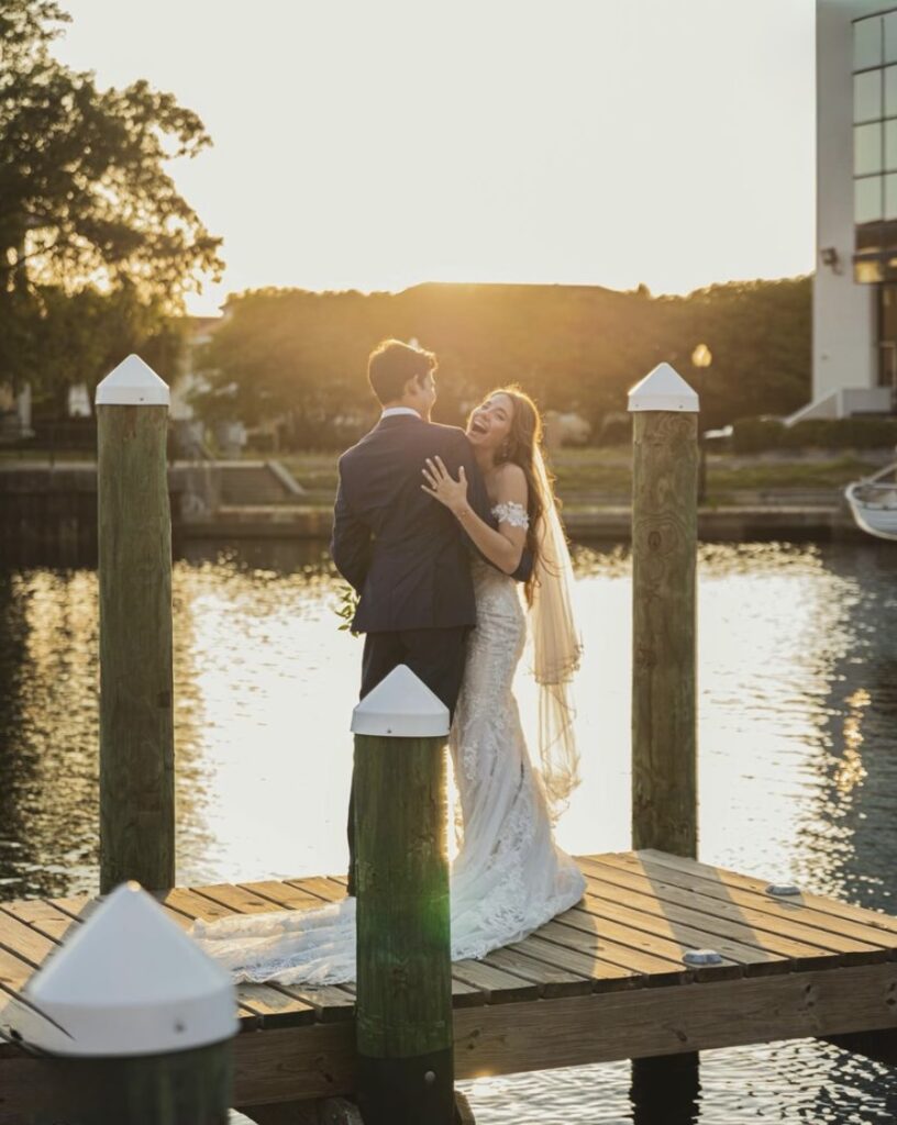 Pensacola Wedding Venues with just married joyful couple on dock at Palafox Wharf Waterfront during golden hour.  Bride with long flowing hair is smiling in a beautiful long wedding dress and veil. https://www.phocusonme.com/photography