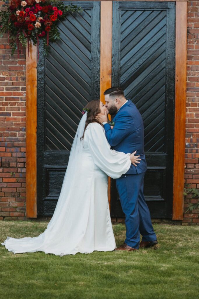 Catie + TJ in front of the iconic black doors to Palafox Wharf Waterfront Venue with a puff of deep red florals during ceremony.  
 What a Stunning Fall Wedding. 
 Photo by Hayle Wilson Photography