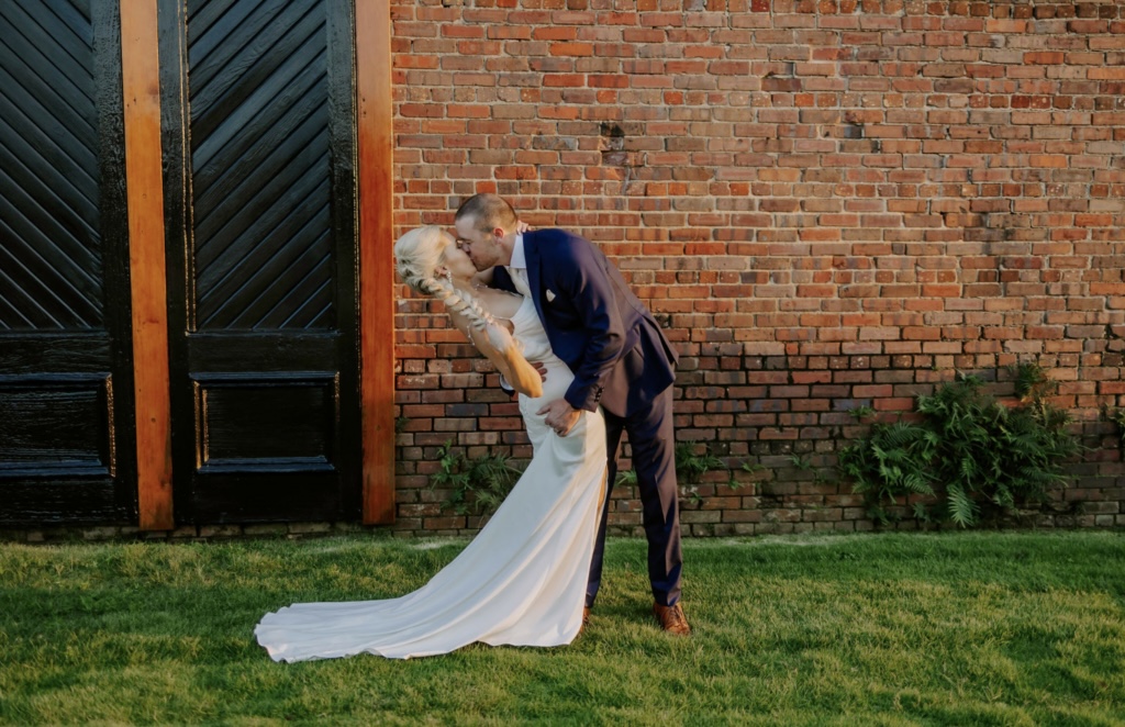 Palafox Wharf Waterfront Weddings and Events Venue with just married couple in the deep embrace on the lush green waterfront lawn in front of the red brick and iconic black doors. Looking gorgeous in her white wedding dress and his dark blue suit. Great example of how to pose for engagement photos.  Photo by Love is Wild Photography.  How to Wedding Plan