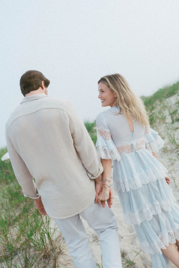 Palafox Wharf Waterfront Wedding and Event Venue showing you this lovely just engaged couple with her in a baby blue lace tiered dress and he is in a light tan linen pressed shirt and white plants and they are walking away from camera in this pose and Great example of how to pose for unique engagement photos. Great easy How to pose.  How to Wedding Plan