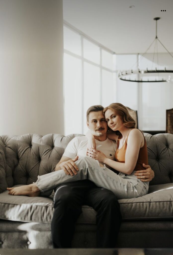 Showing Palafox Wharf Waterfront Wedding Venue with just engaged couple posing with one leaning against the other on a sofa looking into the camera.  Both are casually dressed in jeans and summer wear and Great example of how to pose for unique engagement photos.  How to Wedding Plan