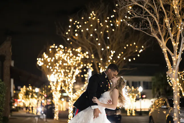 Couple just married at Palafox Wharf Waterfront Wedding Venue with twinkling street lights in the background and she is in a white dress and he is in his military uniform