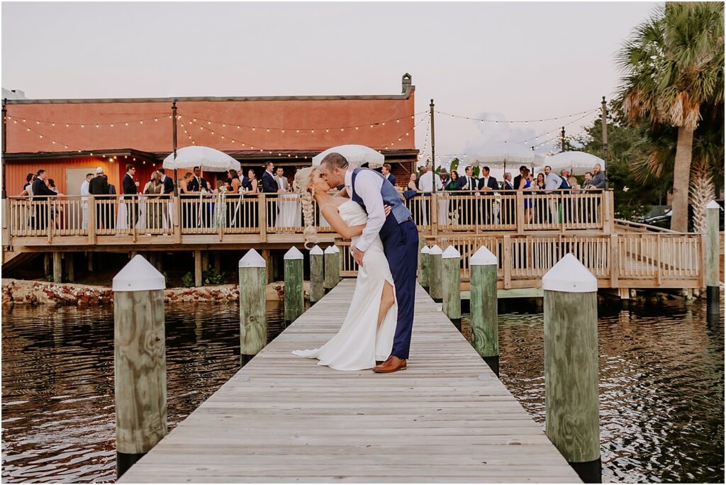 Which is the cheapest place for a destination wedding and the answer is Palafox Wharf Waterfront Wedding Venue and here showing a bride and groom embracing on the waterfront deck with the celebration going on in the background for cocktail hour with 135 guests and cream umbrellas