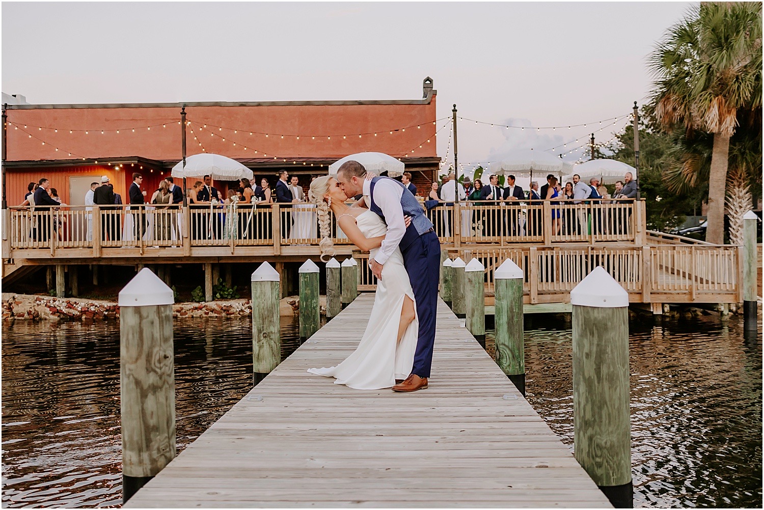 Pensacola wedding venues on a budget featuring Palafox Wharf Waterfront Wedding Venue with Bride and Groom on dock and in the background 135 guests enjoying cocktail hour with bistro tables and guest tables along with fringed cream umbrellas