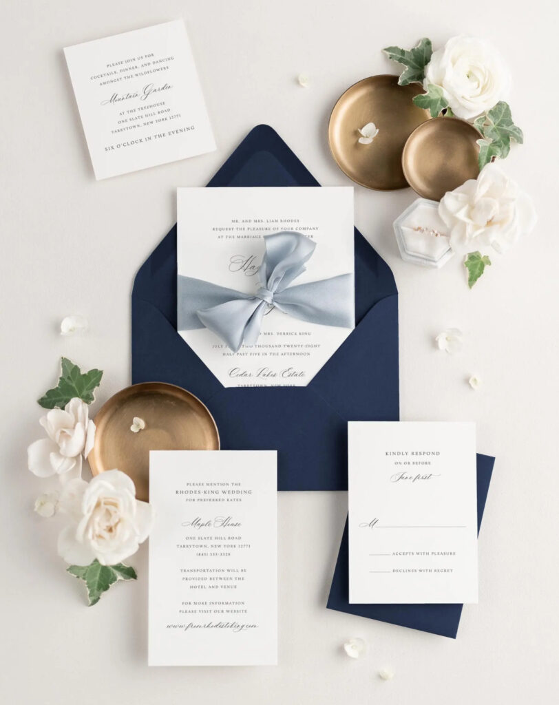 Small wedding venues in Pensacola, Florida which would be Palafox Wharf Waterfront Wedding Venue here showing a beautiful invitation suite flat lay with bronze trays white flowers, engagement ring in box, and greenery and light blue bow and navy envelope 