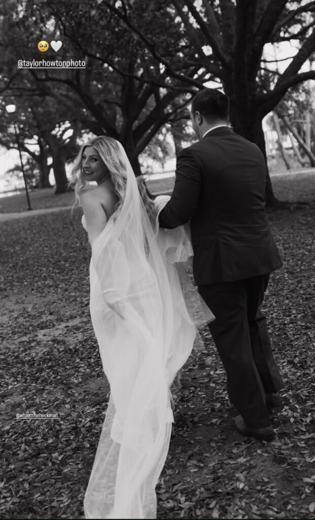 small wedding venues in pensacola, florida and in this case this is a Palafox Wharf Waterfront couple in a black and white photo for pre-wedding photos in a nearby park with bride in her white wedding dress looking back over her shoulder to the photographer and the groom holding part of the veil and he is in his dark suit with big tress in the background and leaves on the ground