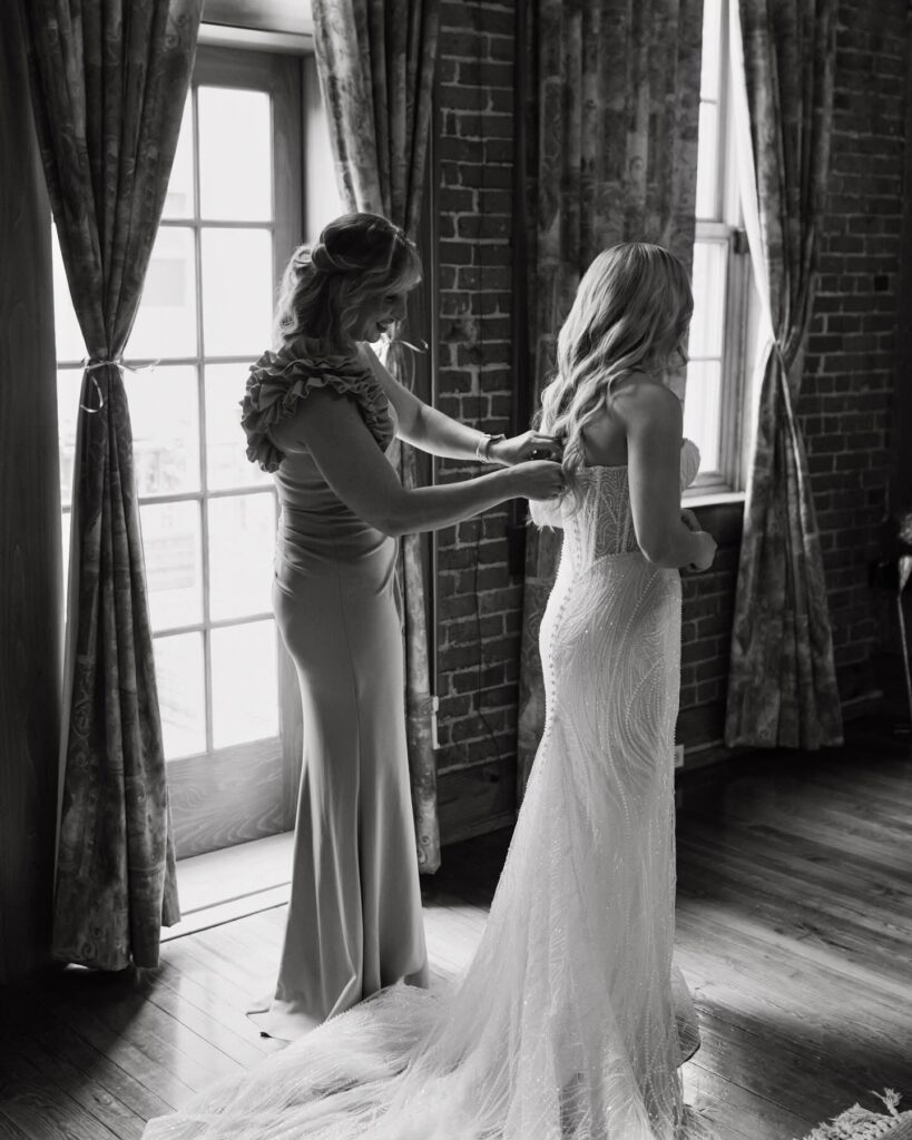 Pensacola wedding venue prices and here is our Palafox Wharf bride and her mom at their get ready accommodations and mom is helping bride put on her wedding dress by buttoning up the back.  This is a back and white photo.