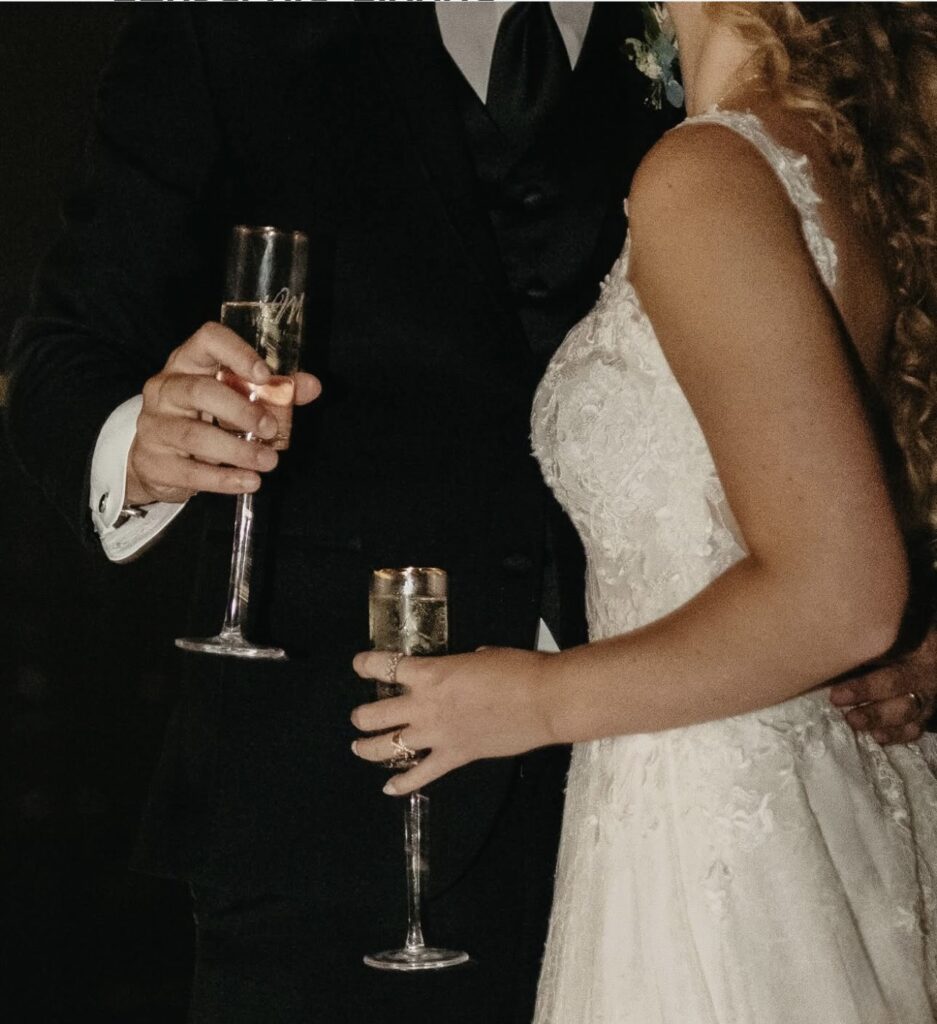 Pensacola wedding venues on a budget with Palafox Wharf Waterfront Wedding Venue showing couple with engraved champagne glasses at their wedding and she is in white lace dress and he is in black suit with cuff links and black tie.