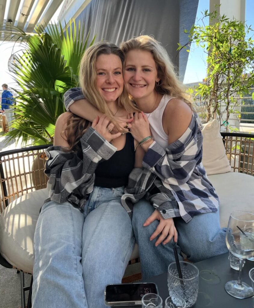 Pensacola wedding venues showing Palafox Wharf Waterfront Wedding Venue’s Bride on the right with her friend in Charleston SC for her engagement sitting on an outdoor white cushioned seat for two with greenery in the background