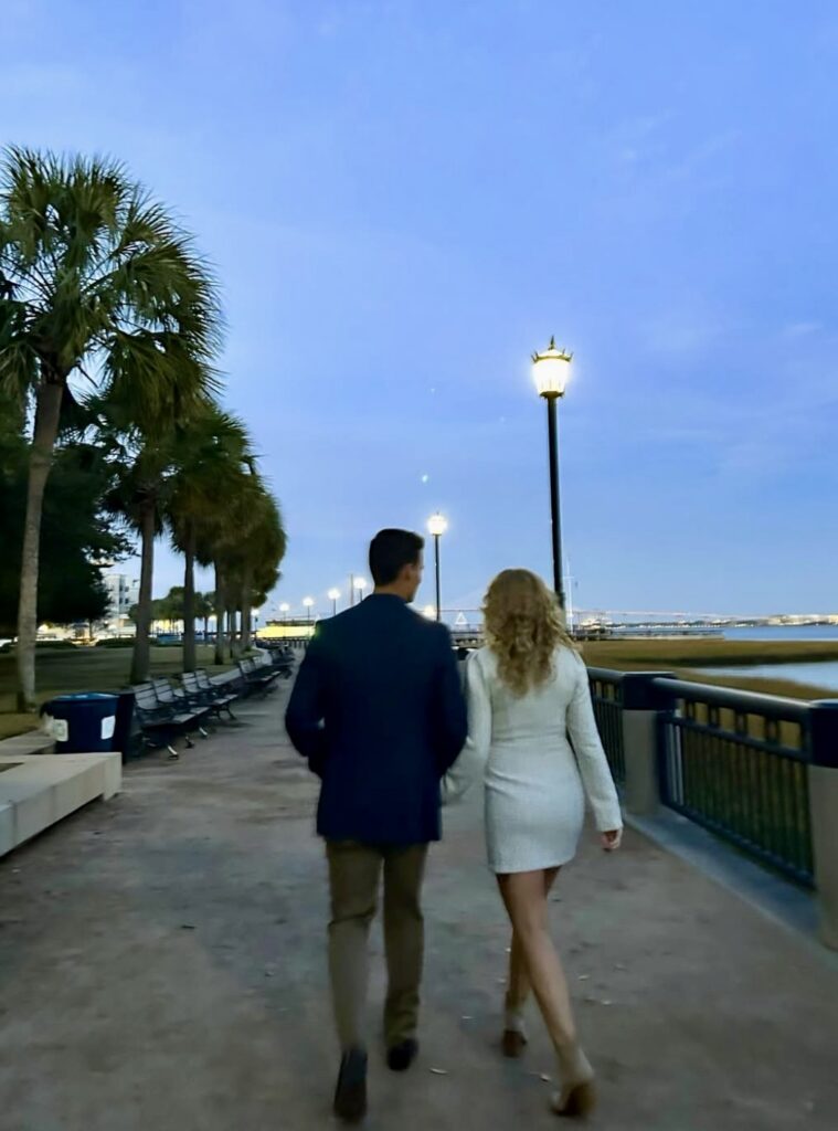 Pensacola Wedding with Palafox Wharf Waterfront’s couple after they were just engaged in Charleston with them at a park in the eventing and she is in a short white long sleeved dress and he is in a blue jacket and khaki pants and they are walk down a promanade