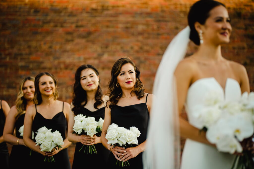 Engaged Couples Can't Get Enough of this Dreamy Southern Downtown Waterfront Wedding Venue Here in Pensacola Fl is Palafox Wharf Waterfront Wedding Venue showing beautiful Amy Bride wearing a sleek white dress and carrying white orchards and in the background are bridesmaids dressed in black with white flowers