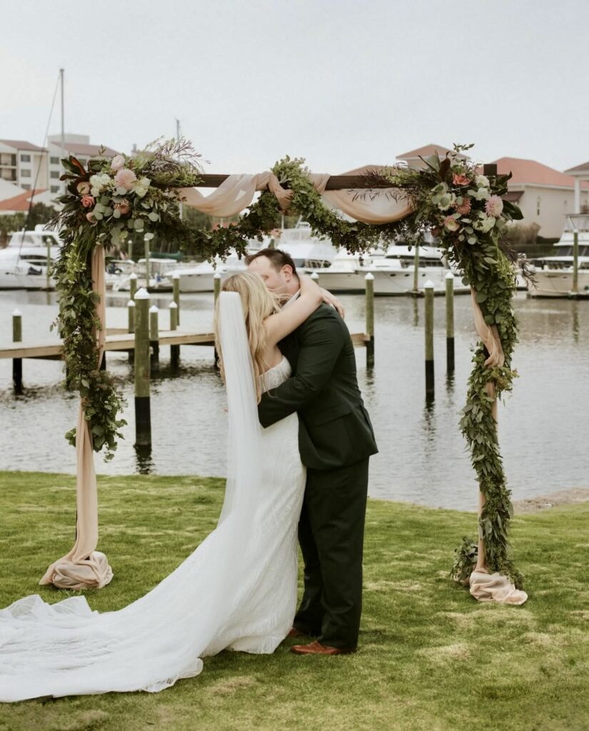 wedding venue pensacola showing couple at Palafox Wharf Waterfront Wedding Venue in front of their wedding arbor adorned with greenery, florals and drapery on green grass with the waterfront in the background