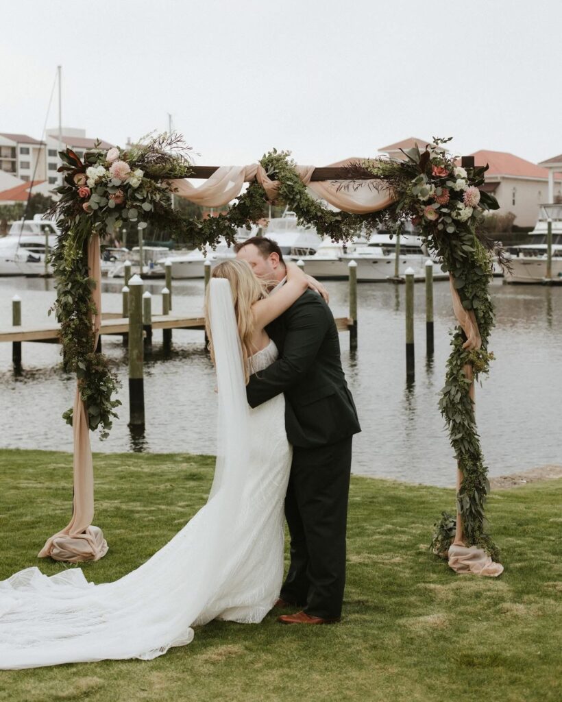 Wedding Venue Pensacola showing just married couple under their arbor with greenery and florals with water in the background an with boats