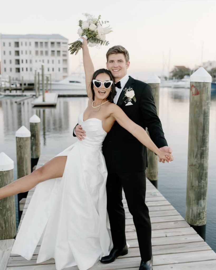 Wedding Venues Pensacola showing their bride and groom just married on the dock with bride in heart shaped sun glasses wearing a white dress and groom in black suite with white shirt and a black bow tie and there are boats docked in the background with a white building also in the background