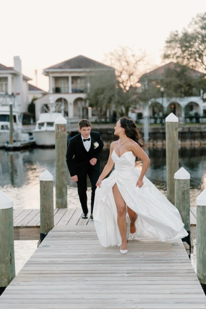 Bride and groom playfully running toward each other on a wooden dock by the water, with boats and waterfront homes in the background
