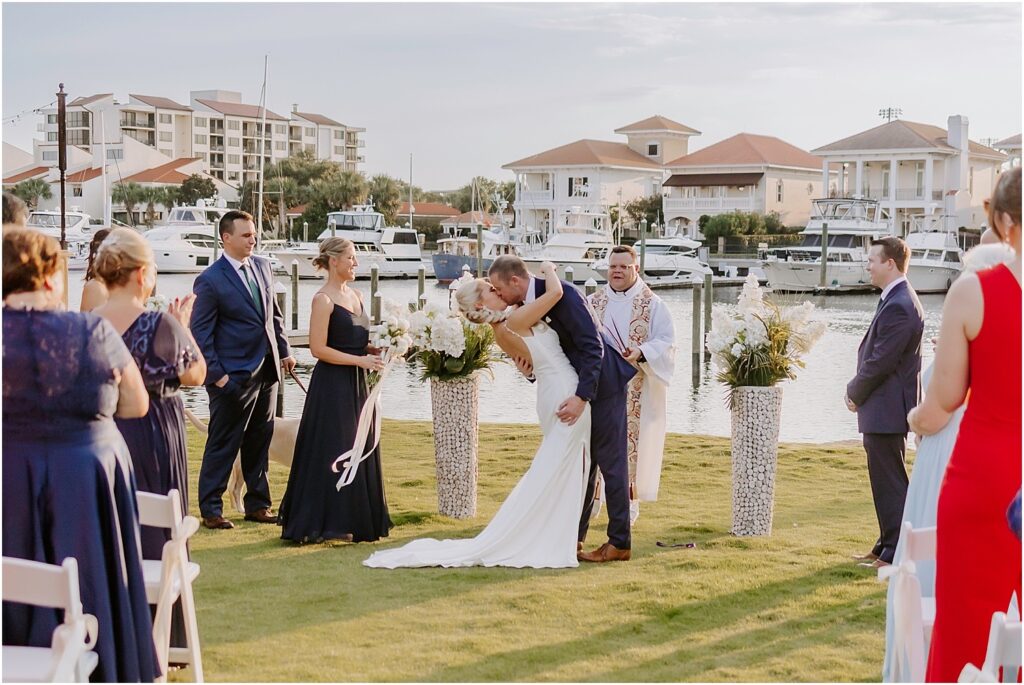 Groom kissing bride at their wedding ceremony on the green at palafox Wharf Waterfront with water and luxury homes in the background