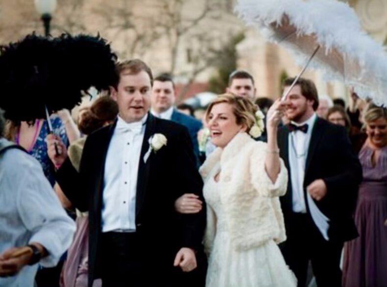 Bride and groom celebrate with a lively New Orleans-style second line parade during their downtown Pensacola wedding at Palafox Wharf Waterfront. The bride holds a white feathered parasol while smiling guests follow behind with festive energy.
