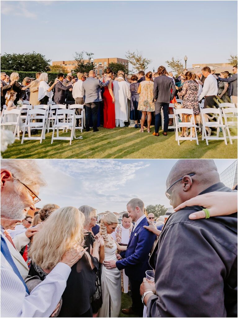 Unforgettable wedding guest experience as they gather in prayer around the bride and groom during an outdoor ceremony on the lawn at Palafox Wharf Waterfront in downtown Pensacola. The couple is surrounded by loved ones in a heartfelt moment of blessing under clear blue skies.
