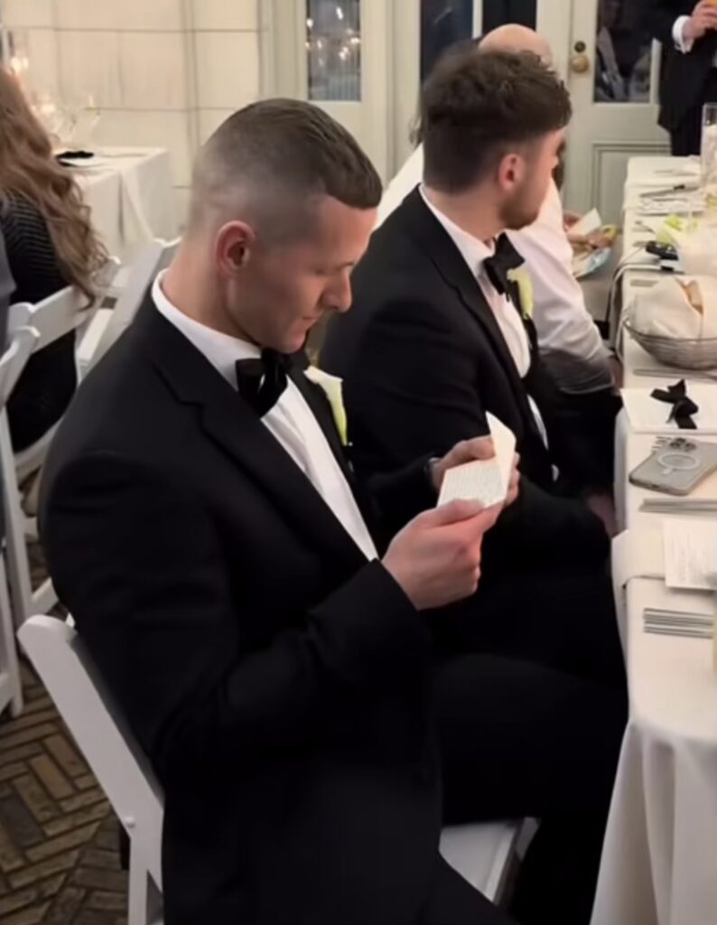 A man dressed in a tuxedo reads a book, surrounded by others seated at tables in a lively atmosphere.