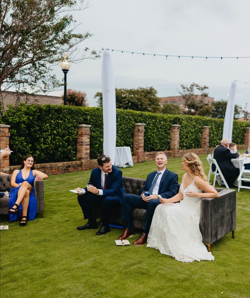 Bride seated on dark gray lounge furniture chatting with guests which enjoying a drink and a bit to eat on the waterfront lawn with hedge behind them at Palafox Wharf Waterfront Wedding Venue Pensacola Florida