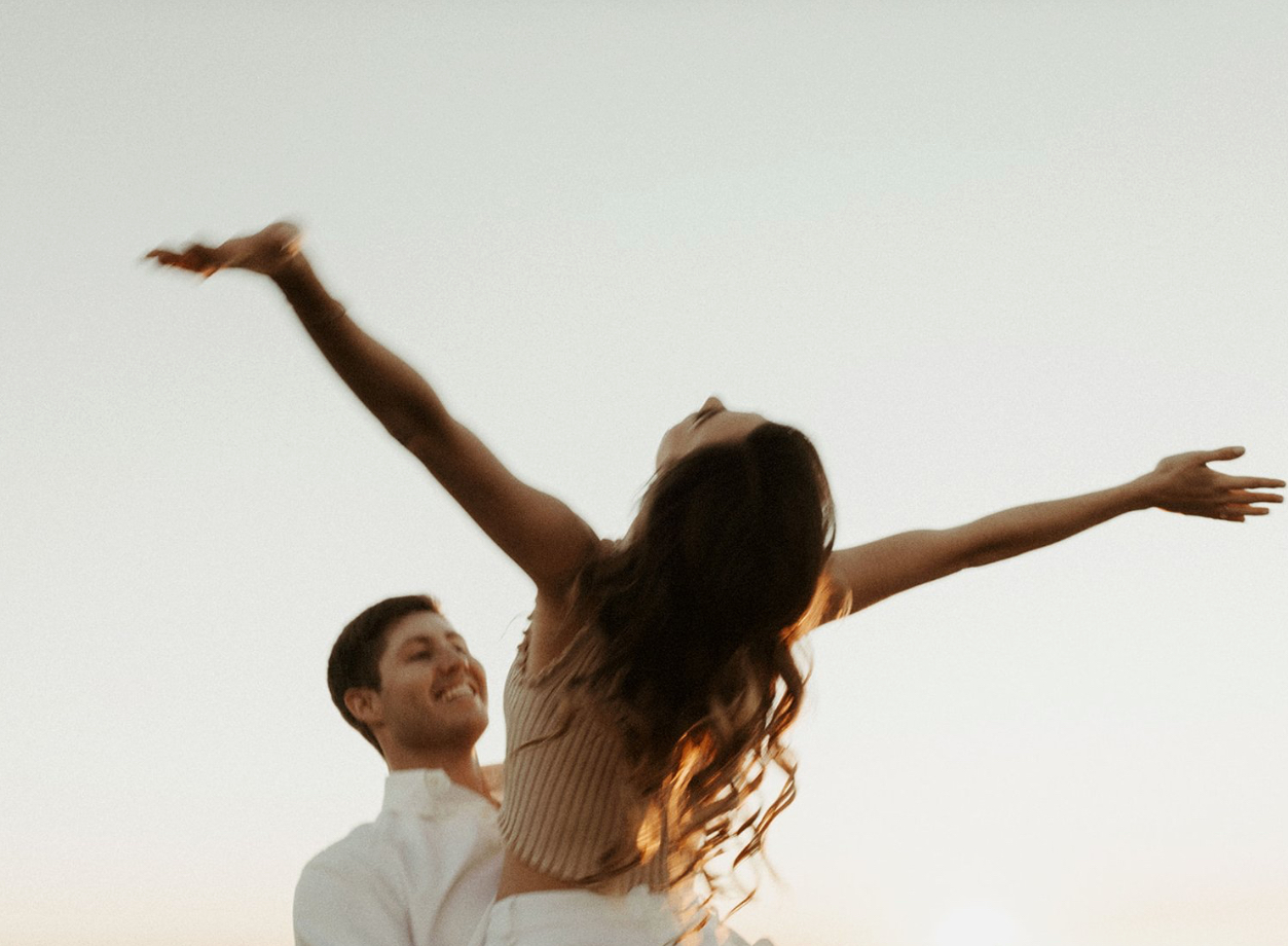 Romantic sunset engagement photo of a joyful couple at a Gulf Coast state park. Woman with arms outstretched in carefree celebration as man lifts her, glowing in golden light. Perfect for wedding proposal inspiration near Pensacola or Florida beaches.