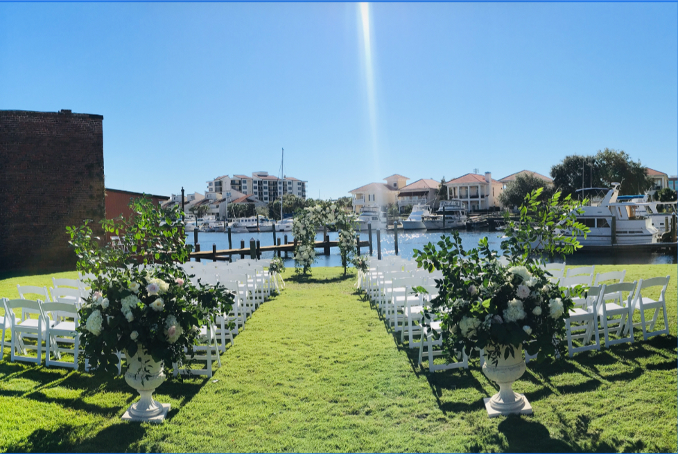Pensacola Wedding Venue showing Waterfront wedding ceremony setup at Palafox Wharf with white chairs aligned in rows on lush green grass, floral arrangements in white urns, and a floral arbor facing the dock and marina under a bright blue sky. Wedding Ceremony Rehearsal
