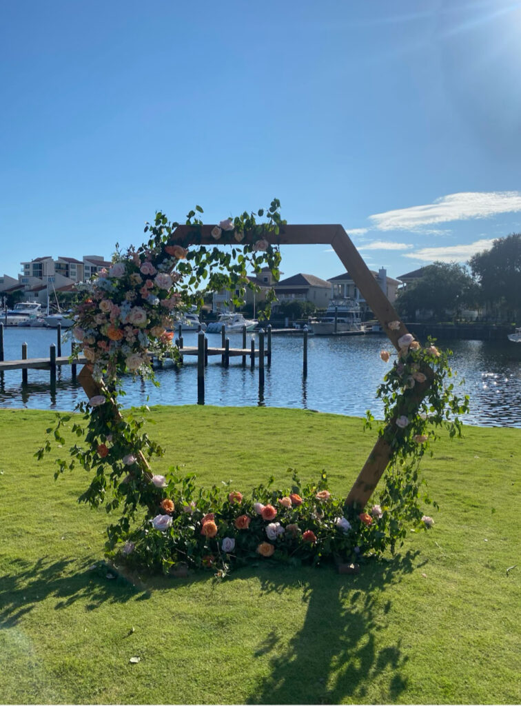 wedding venues pensacola fl with a beautifully octagon ceremony wedding arbor on the green lawn that feels like a carpet with pretty florals on the arbor set with the blue water in the background with boats and upscale homes
