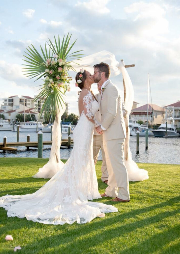 the palafox wharf waterfront wedding venue Pensacola with a bride and groom kissing in front of a tropical floral arrangement on a draped arbor on the great lawn and in front of the blue waterfront at palafox Wharf Waterfront wedding venue