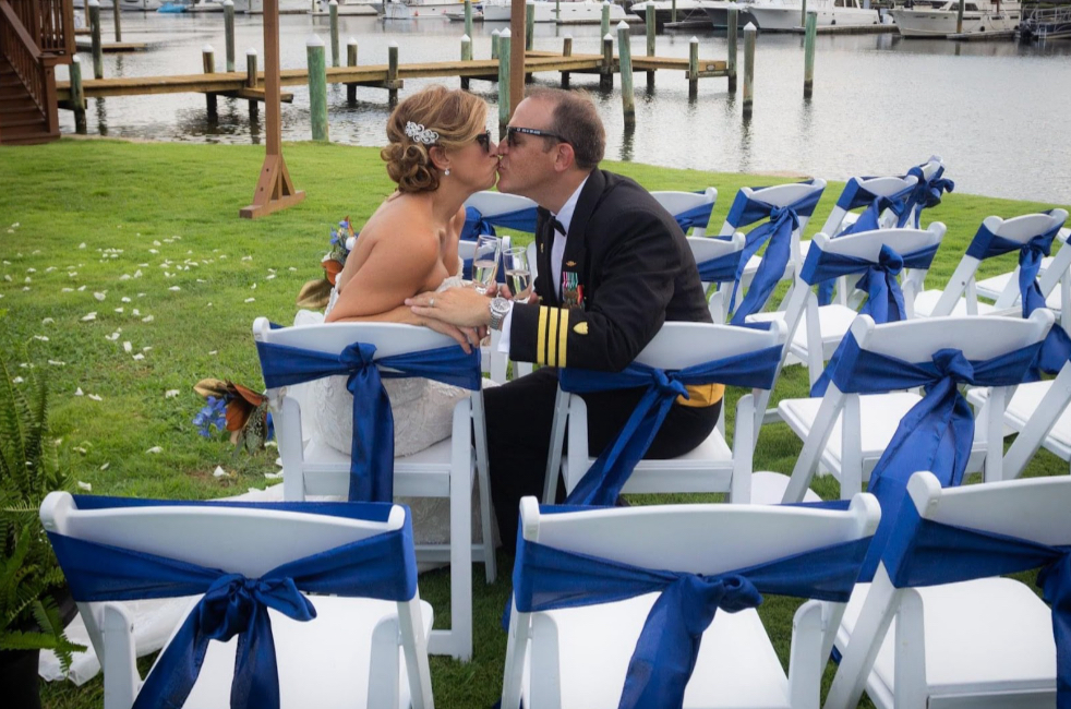 Palafox Wharf Waterfront Wedding Venue’s military United States Coast Guard Commander marrying a head of a Government agency sitting on their ceremony chairs with the sunglasses on kissing on the green verdant lawn with the water and dock in the background
