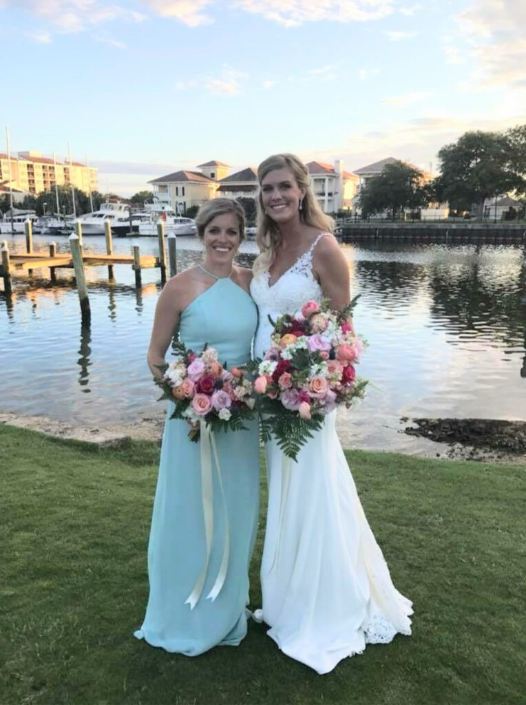 Palafox wharf waterfront wedding venue photos with with the bride and her maid of honor in light blue with beautiful bouquets of pink, red, green, and blush standing on the green lawn by the water with dock, boats and homes in the back ground along with large trees
