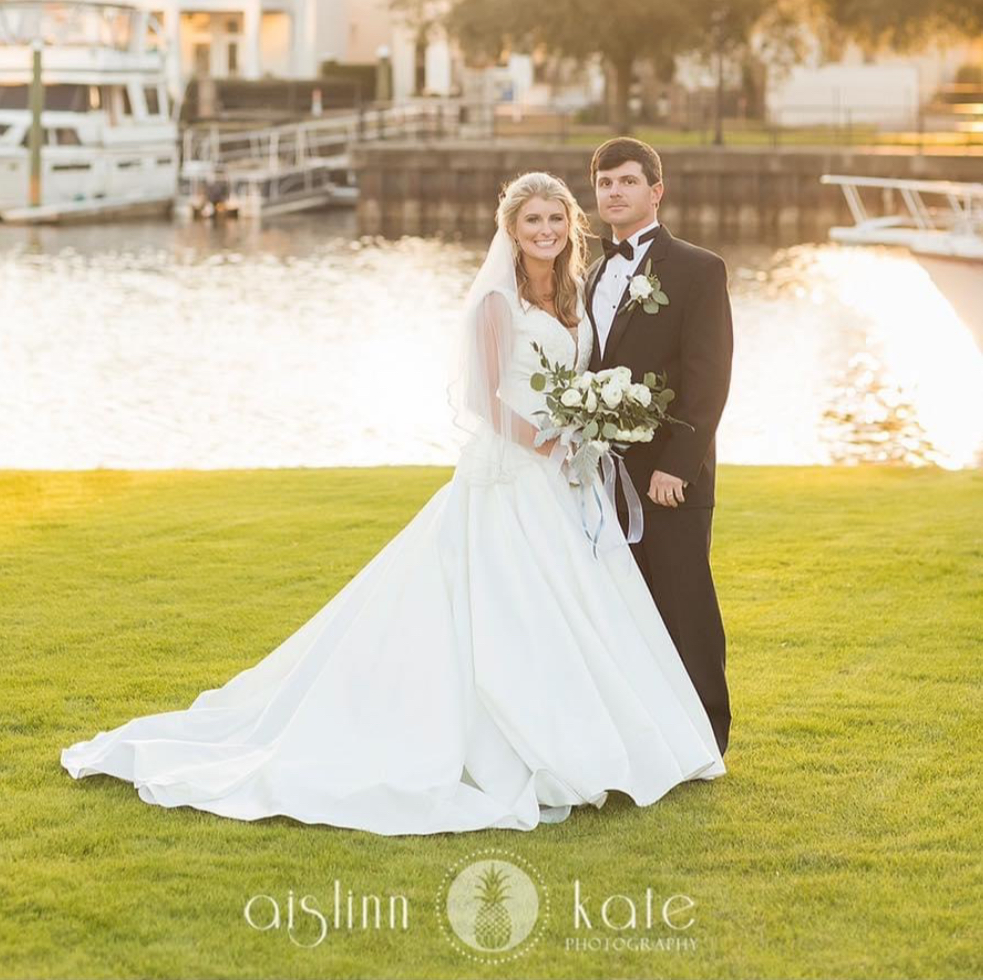 palafox wharf waterfront wedding venue with a bride and groom in formal wear for their wedding and she is in white with a veil and blond hair and he is in black with a black bow tie standing on the gorgeous green grass with the water, boats, and sun shining down on the water
