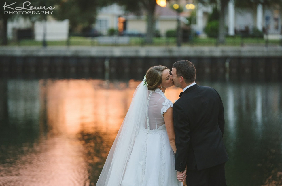 wedding venues pensacola with bride and groom in front of the water with the golden light from the sun shining down on the water and they are kissing and she has a long veil with a white dress that belonged to her mother who was married in that dress