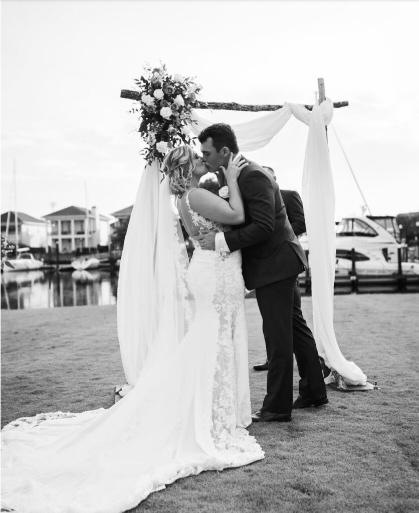 Wedding venues pensacola fl with black and white photo of bride and groom kissing in front of their hand crafted arbor which has florals and is draped nicely with white drapery.  They are on the lawn with the water, boats, and homes in the background
