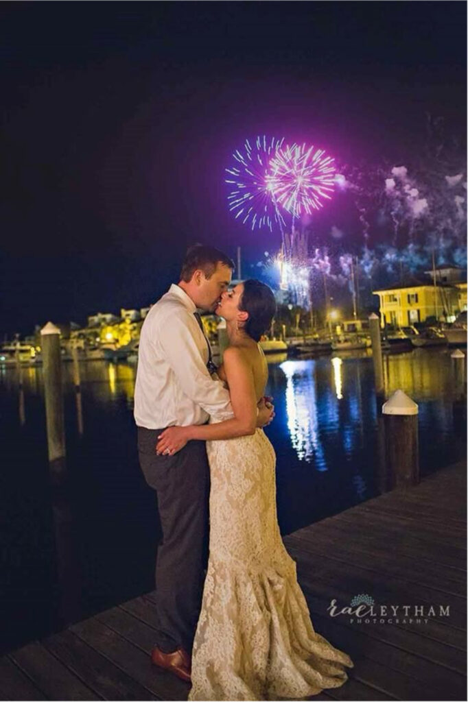 Palafox wharf waterfront wedding venue with very nice fireworks with the bride and groom kissing and she is in a soft beige wedding dress and he is in black pants, white shirt and tie with both standing on the waterfront dock
