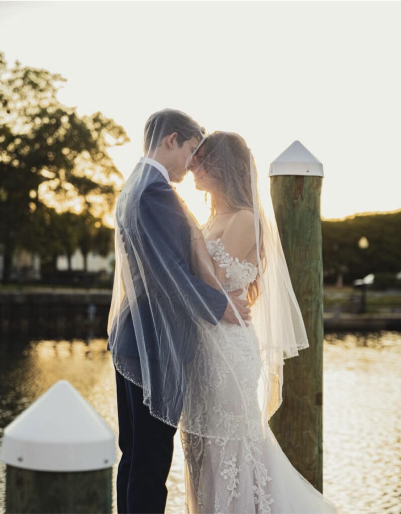 Wedding places in pensacola fl with the bride and groom both under her veil during the golden hour on the dock with the water in the background.  She is in an off the shoulder dress in white and he is a blue suit.
