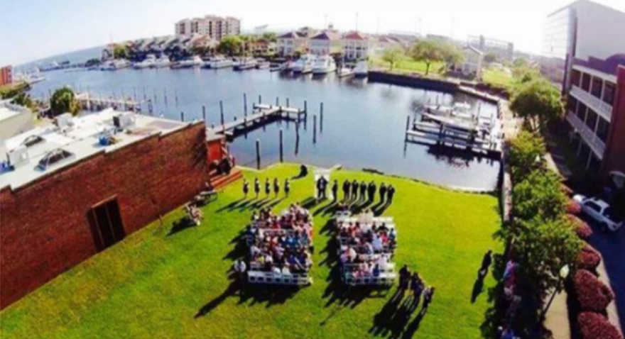 Pensacola wedding venues with the waterfront wedding venue called Palafox Wharf Waterfront Wedding Venue here with a bird’s eye view of a lawn ceremony on the waterfront with musicians.  This shows the venue to the left, the water with boats and homes.