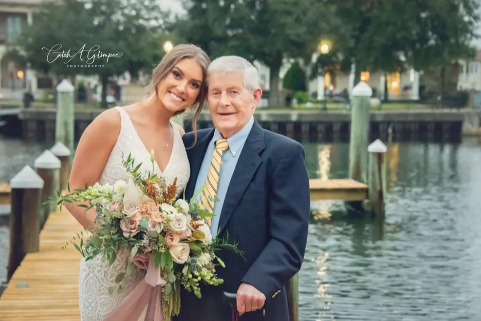 Pretty bride and her grandfather in front of the dock and water with her holding her large bridal bouquet and he has a cane.  He is in blue suit with yellow striped tie.