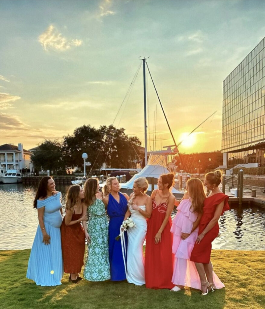 Pensacola wedding sites here at Palafox Wharf Waterfront wedding venue showing the golden hour with the late afternoon sun, boats, building, homes, trees in the back and in the foreground , the beautiful bride with her bridesmaids all in difference dolor and patterned dressed.  All very chic.
