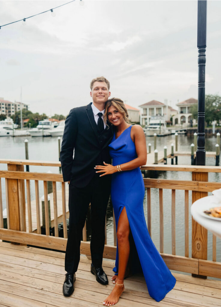 waterfront wedding venues near me with this being Palafox Wharf Waterfront Wedding Venue with bridesmaid dressed in royal blue and husband in black suit, black tie, and black shoes.  They are on the deck at the venue with the water behind them with homes and boats.
