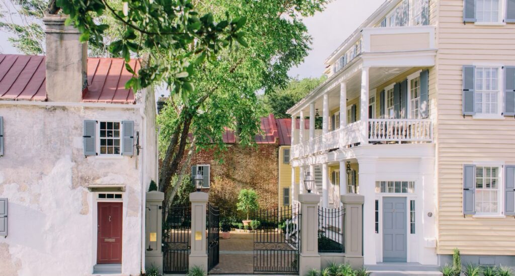 This charming photo showcases a picturesque scene featuring historic architecture with a blend of rustic and elegant elements, perfect for a special occasion. The image captures a quaint street lined with well-preserved buildings, including a weathered stone house with a striking red door and a pastel yellow structure adorned with a white balcony and blue shutters. Lush green trees frame the view, adding a touch of nature, while a wrought-iron gate opens to a serene courtyard where the newly engaged couple are talking about the inviting ambiance of the palafox wharf waterfront wedding venue in Pensacola, FL. The warm lighting and classic design make it an ideal backdrop for a romantic waterfront wedding, blending historic charm with scenic beauty.
